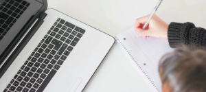 Mujer tomando nota en un cuaderno al frente de un computador portátil.
