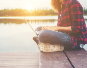 Mujer con un computador portátil en su regazo y sentada frente a un cuerpo de agua mientras el sol despunta en el horizonte.