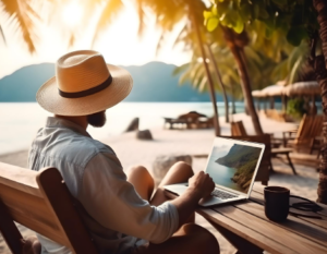Hombre con sombrero, en la playa, disfruta un día de sol mientras interactúa con un computador portátil a la mesa.