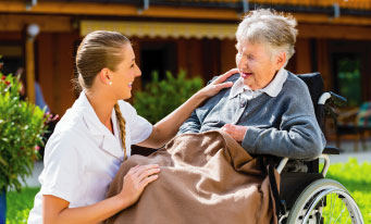 enfermera poniendo su mano sobre el hombro de una mujer mayor que se encuentra sentada en una silla de ruedas.