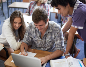 Tres estudiantes universitarios frente a un computador portátil, uno de ellos está escribiendo en una hoja de papel.