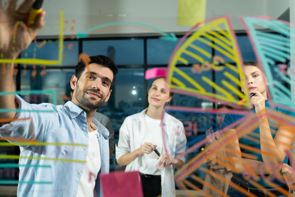 Un hombre y dos mujeres, en actitud de planeación, hacen trazados de colores en un vidrio.
