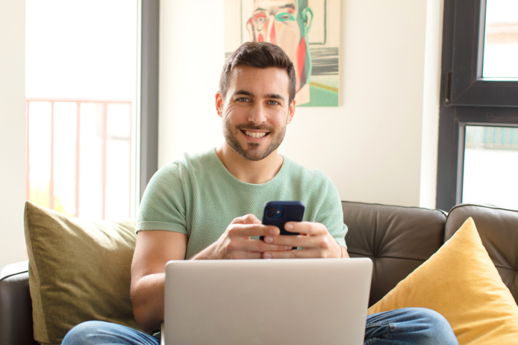 Hombre sentado frente a un computador portátil sosteniendo un celular en sus manos.